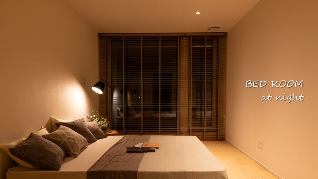 【照明をおさえたおちつく寝室】
バルコニーにも出られる寝室はウッドブラインドをアクセントにシンプルでぬくもりある空間。温かみあるおさえた照明がここちよい眠りを誘う。